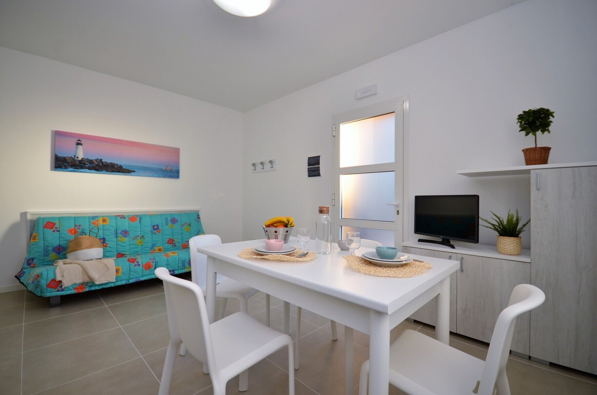 A compact living space with a dining table, white chairs, a sofa with a colorful quilt, a television, and a painting of a lighthouse.
