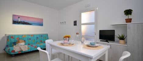 A compact living space with a dining table, white chairs, a sofa with a colorful quilt, a television, and a painting of a lighthouse.