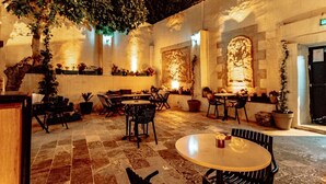 Un espace extérieur aménagé en terrasse, avec un sol en pierre, plusieurs tables et chaises, des plantes en pot et des luminaires fixés au mur.