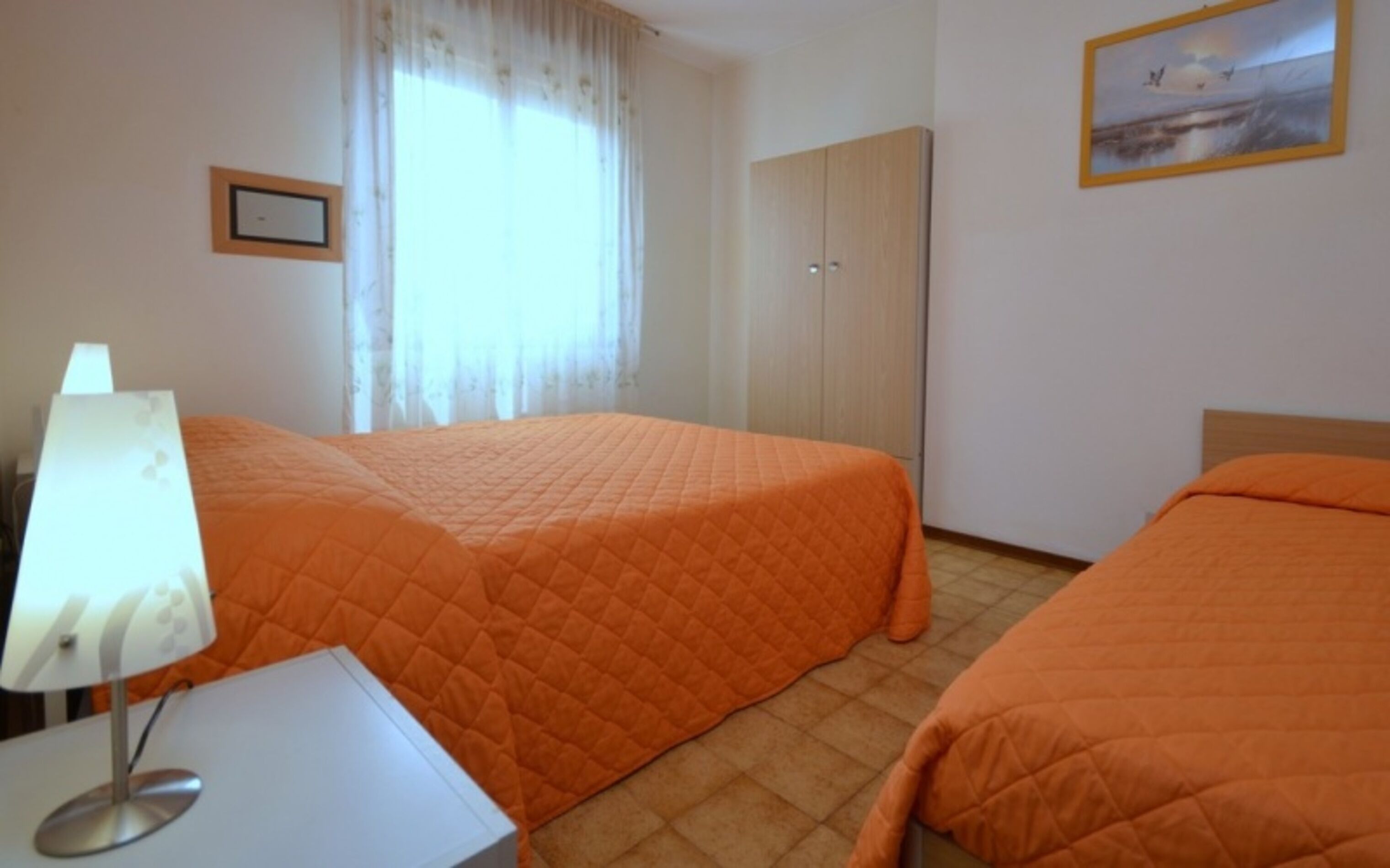 A hotel room with two beds, a desk with a lamp, a wardrobe, and a painting on the wall.