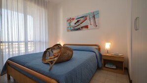 A bedroom with a bed, a nightstand, a lamp, a basket, and a painting of boats on the wall.