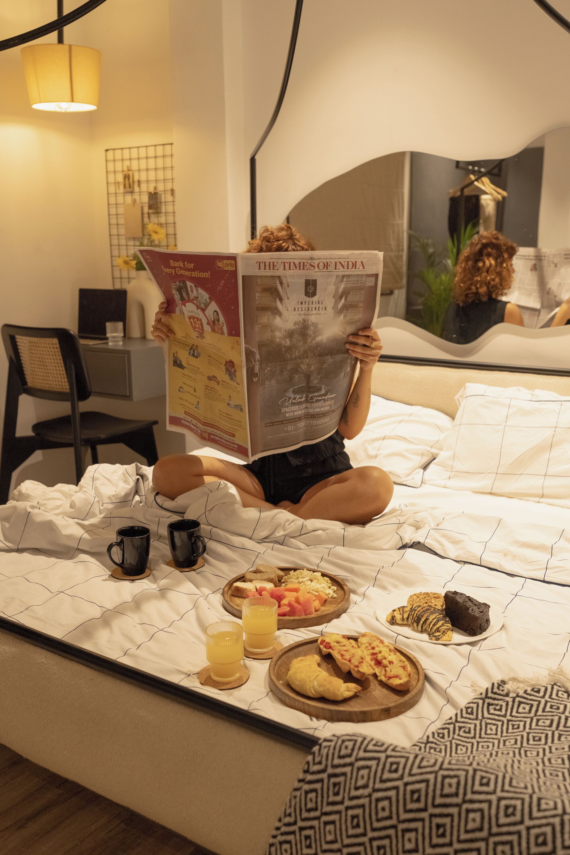A person sitting on a bed reading a newspaper, with breakfast items and two cups on the bed.