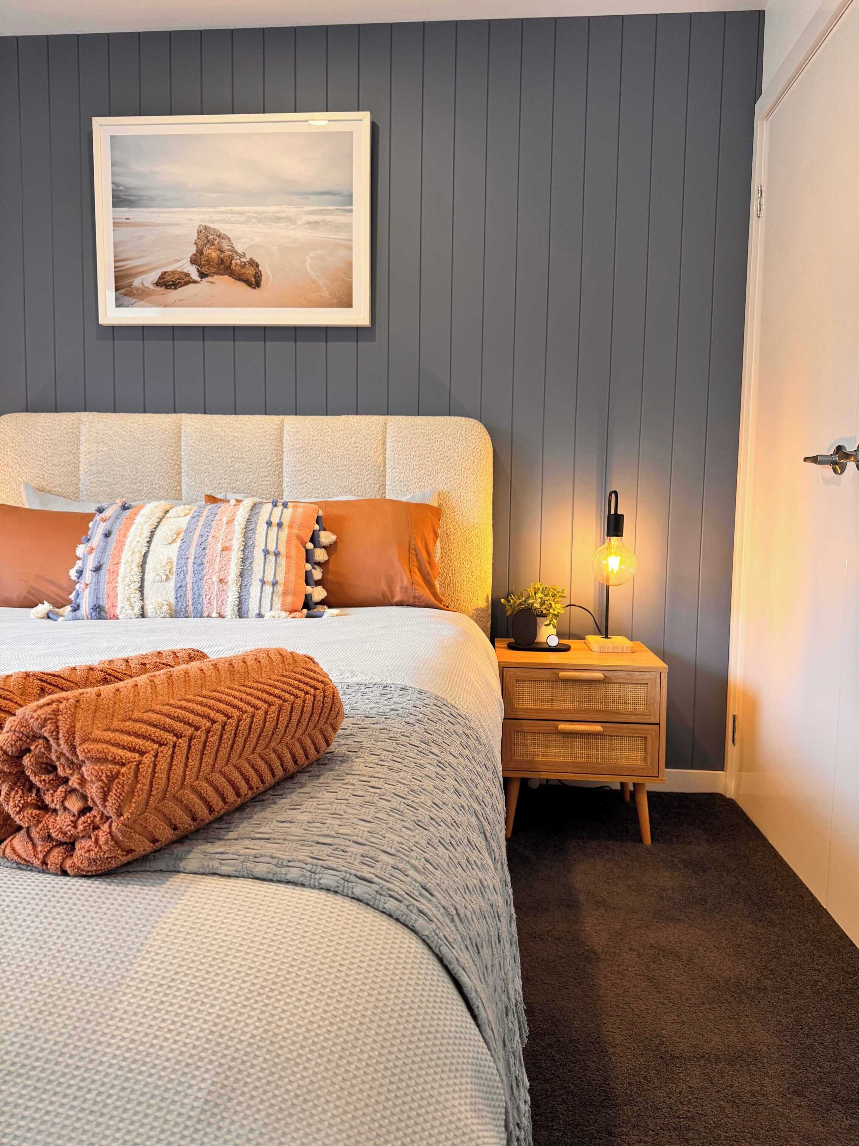 A bedroom with a bed, pillows, a blanket, a bedside table with a lamp, and a framed picture on the wall.