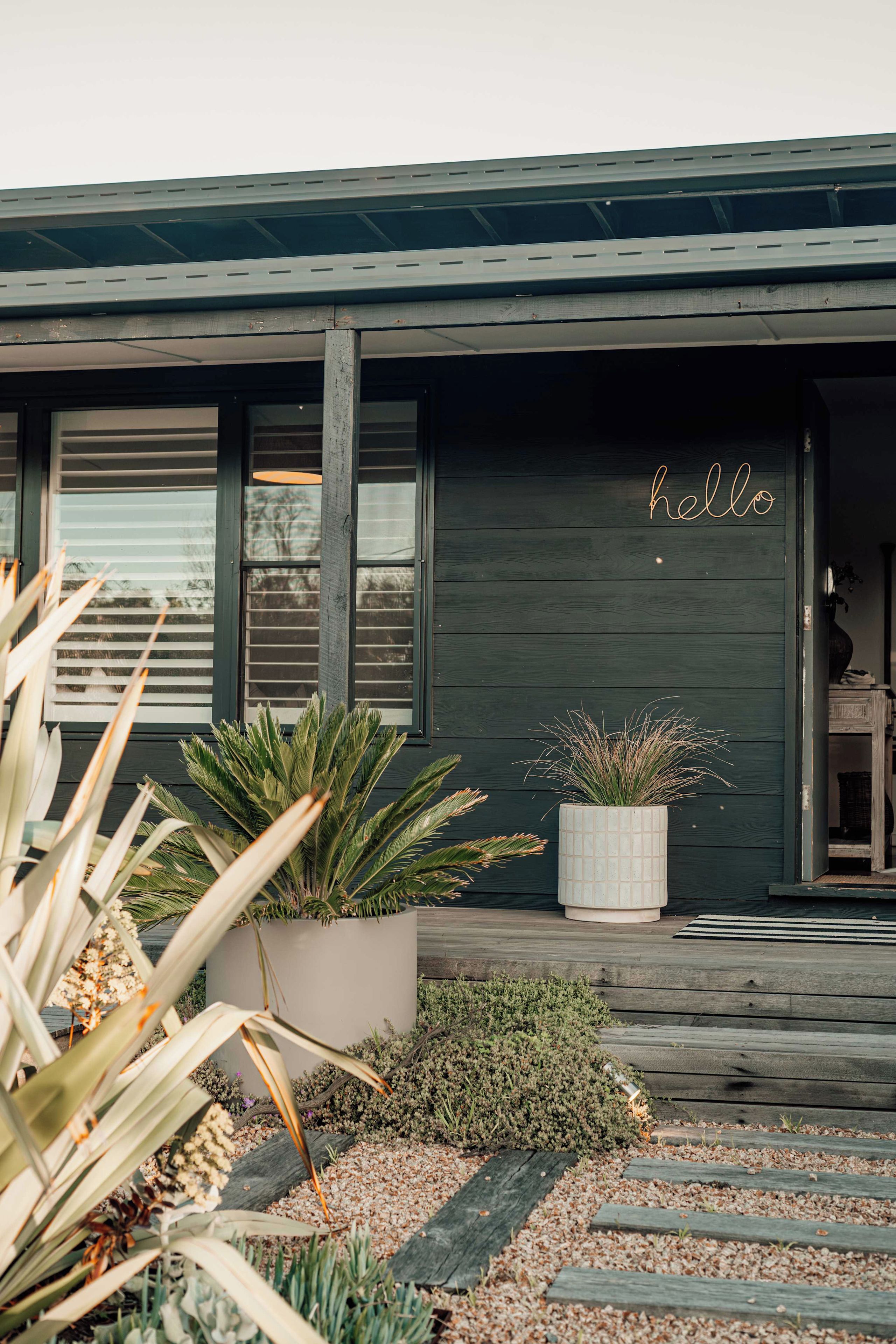 Una casa moderna con facciata scura, un cartello accogliente con la scritta 'hello' e un giardino curato con diverse piante.