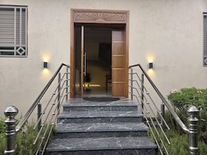 A set of marble stairs leading to a wooden door with a decorative frame, flanked by metal railings and wall-mounted lights.