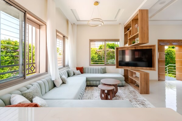 A modern living room with a sectional sofa, a flat-screen TV, and a large window offering a view of greenery.
