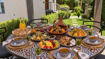 Une table à manger installée en plein air, dressée avec une variété de plats, dont un tajine, des salades et du pain, le tout mis en valeur par un bâtiment et de la verdure en arrière-plan.