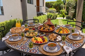 An outdoor dining table with a variety of dishes, including a tagine, salads, and bread, set against a backdrop of a building and greenery.