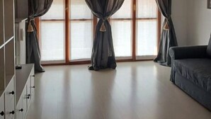 Un salón amplio con un ventanal grande, cortinas oscuras, una araña y un sofá.