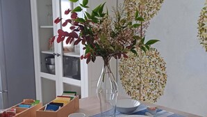 Comedor con una mesa dispuesta para cuatro, incluyendo platos, tazones y cubiertos. Un jarrón con flores está sobre la mesa, y hay cajas de almacenamiento de madera a la izquierda.