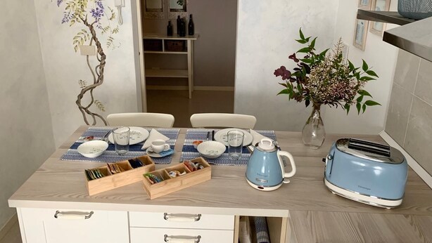 Un comedor con una mesa dispuesta para dos personas, un florero con flores, una tetera y un tostador.