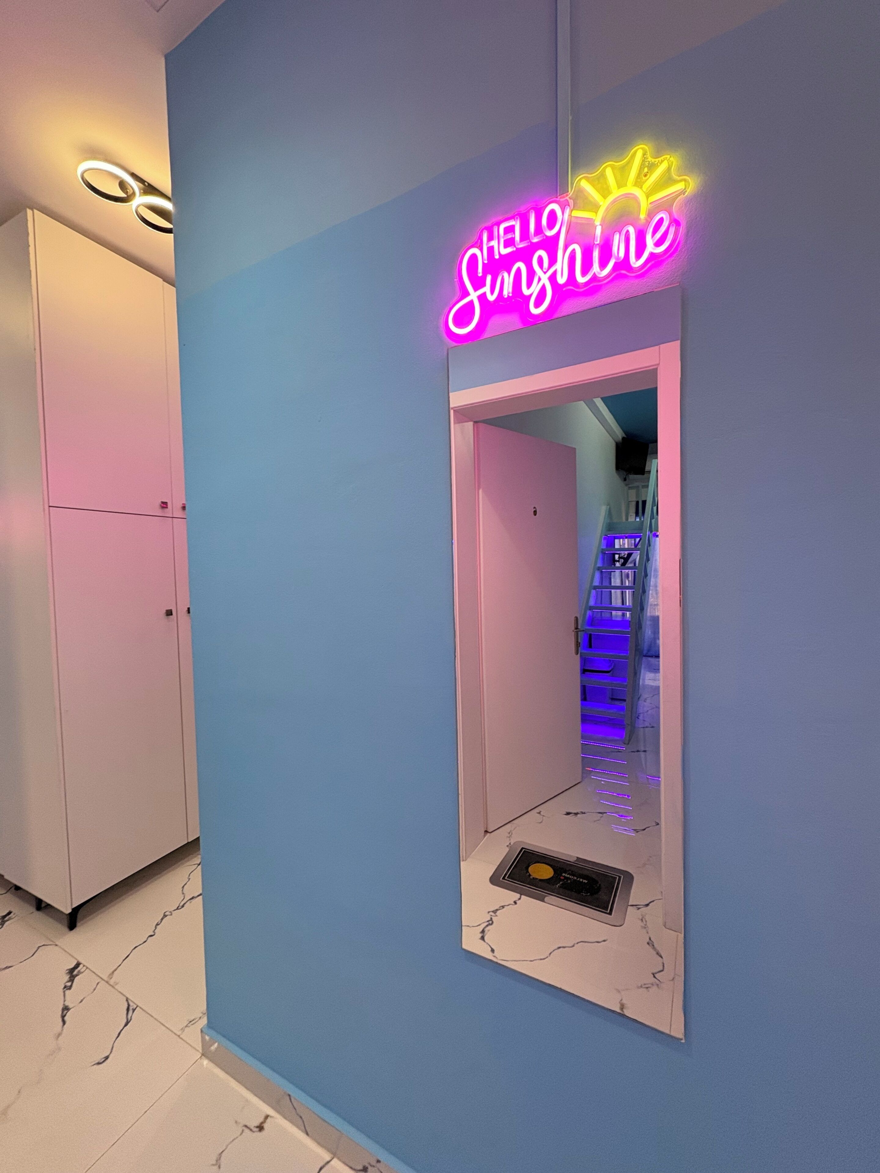 A hallway with a neon sign reading 'HELLO Sunshine' and a staircase with blue lighting.