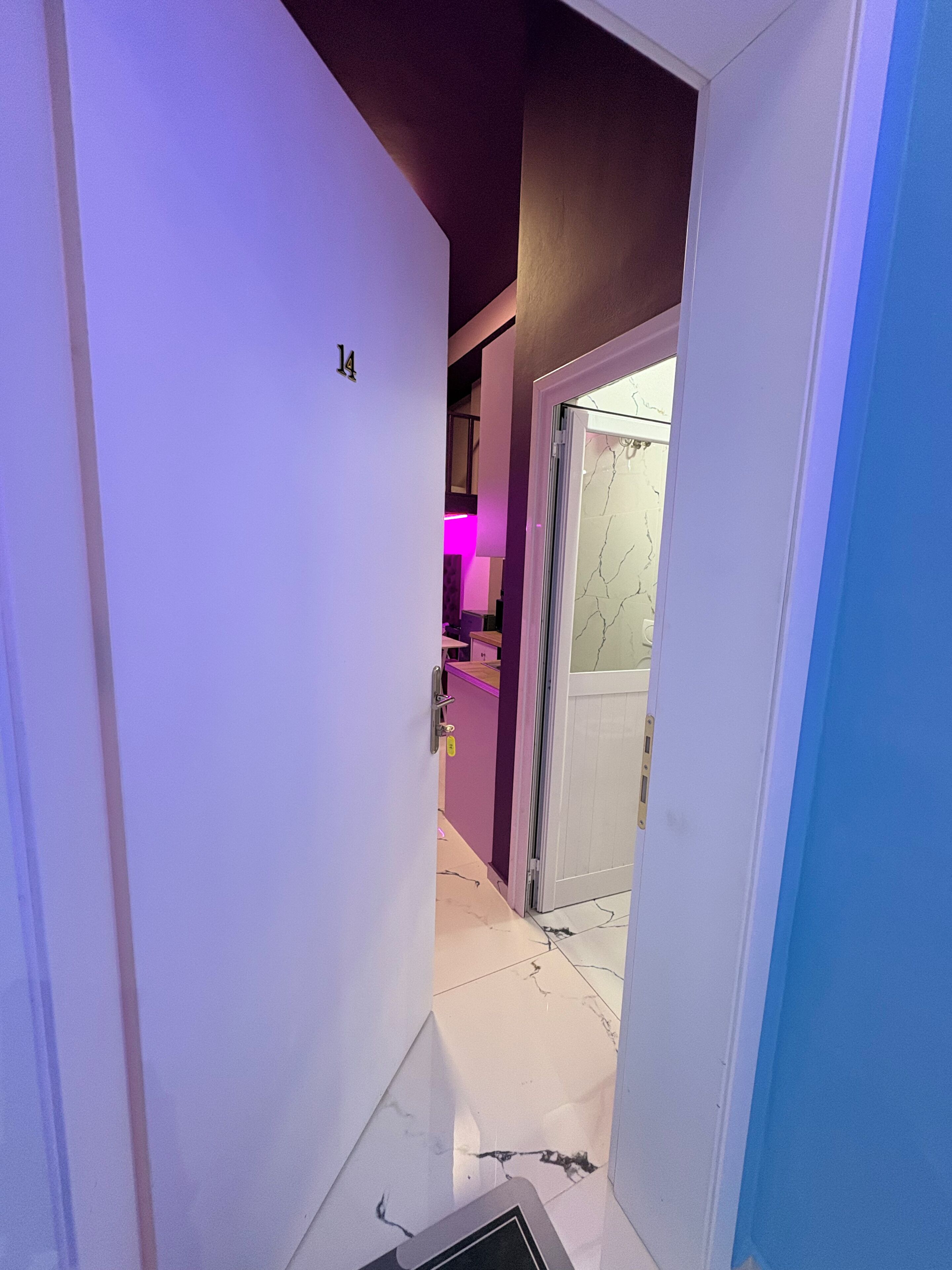 A narrow hallway with a door marked '14' leading to a room with a purple wall and a pink-lit area.