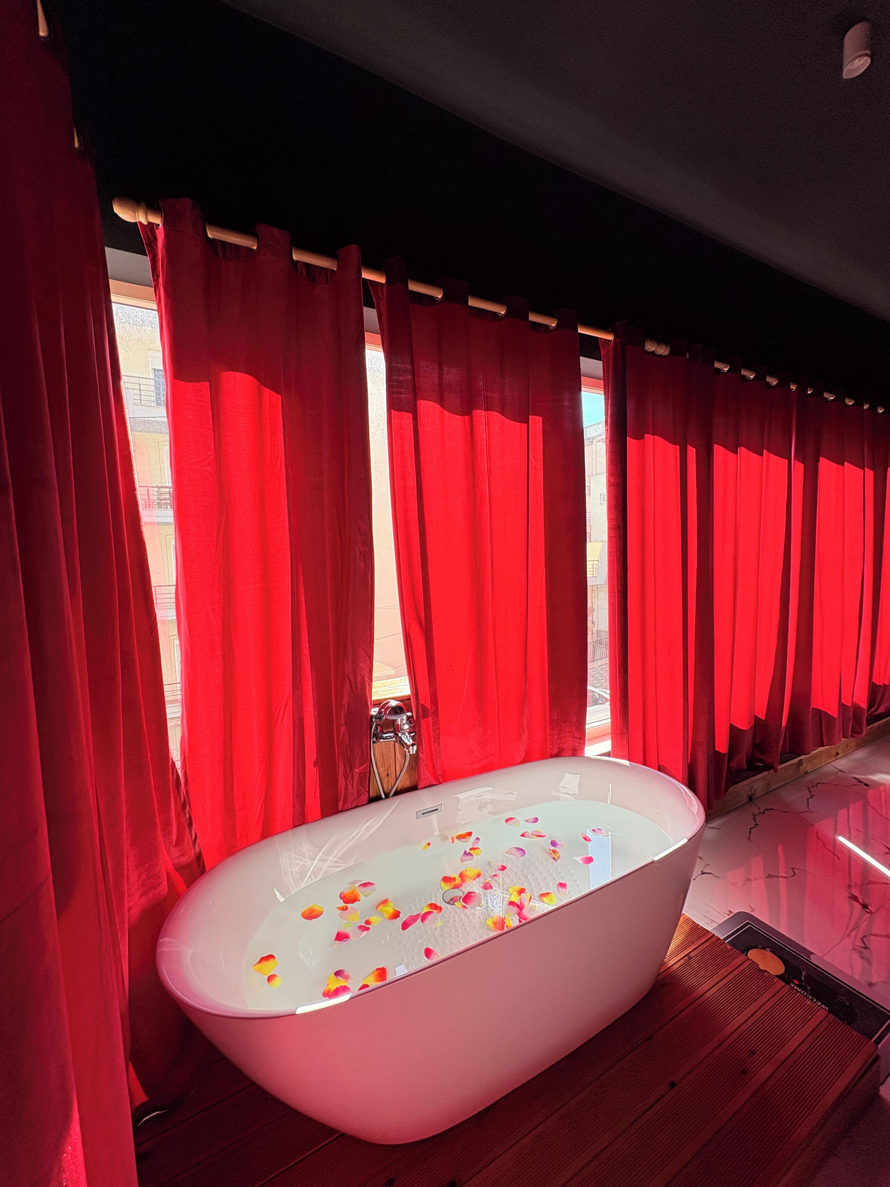 A bathtub with rose petals, red curtains, and a wooden floor.