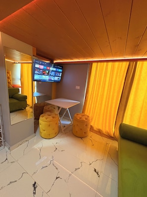 Quarto de hotel com televisão de tela plana presa na parede, uma mesa pequena, dois pufes amarelos, um sofá verde e uma janela com cortinas amarelas.