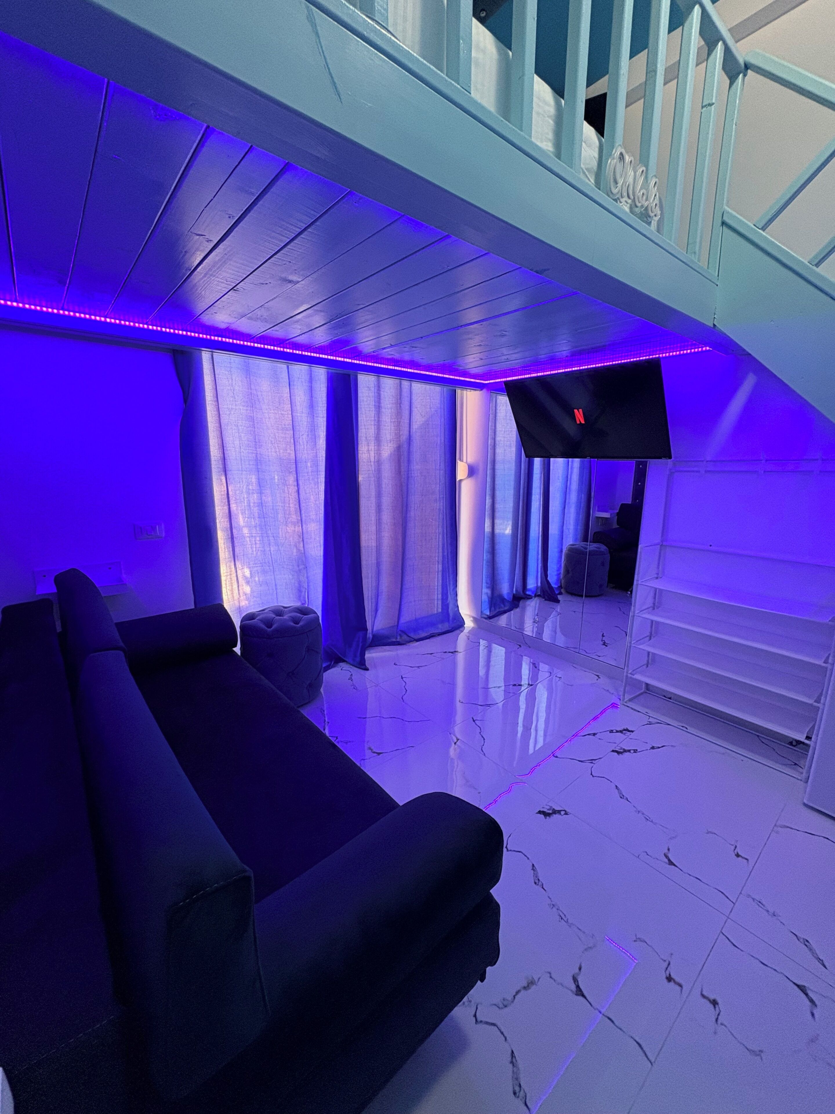 Un salon moderne avec un canapé noir, un téléviseur fixé au mur et un escalier menant à un niveau supérieur.