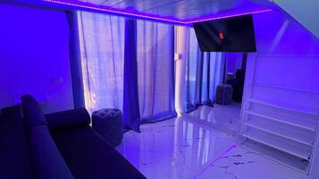 Un salon moderne avec un canapé noir, un téléviseur fixé au mur et un escalier menant à un niveau supérieur.