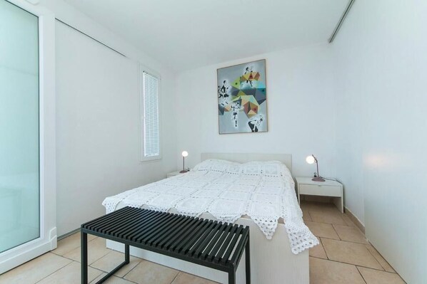 A bedroom with a bed, a bedside table, a nightstand, a bench, a wall art piece, and a window with blinds.