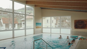 Une piscine intérieure dotée de grandes fenêtres, offrant une vue sur les maisons, et ornée d’une  œuvre d’art en forme de cœur sur le mur.