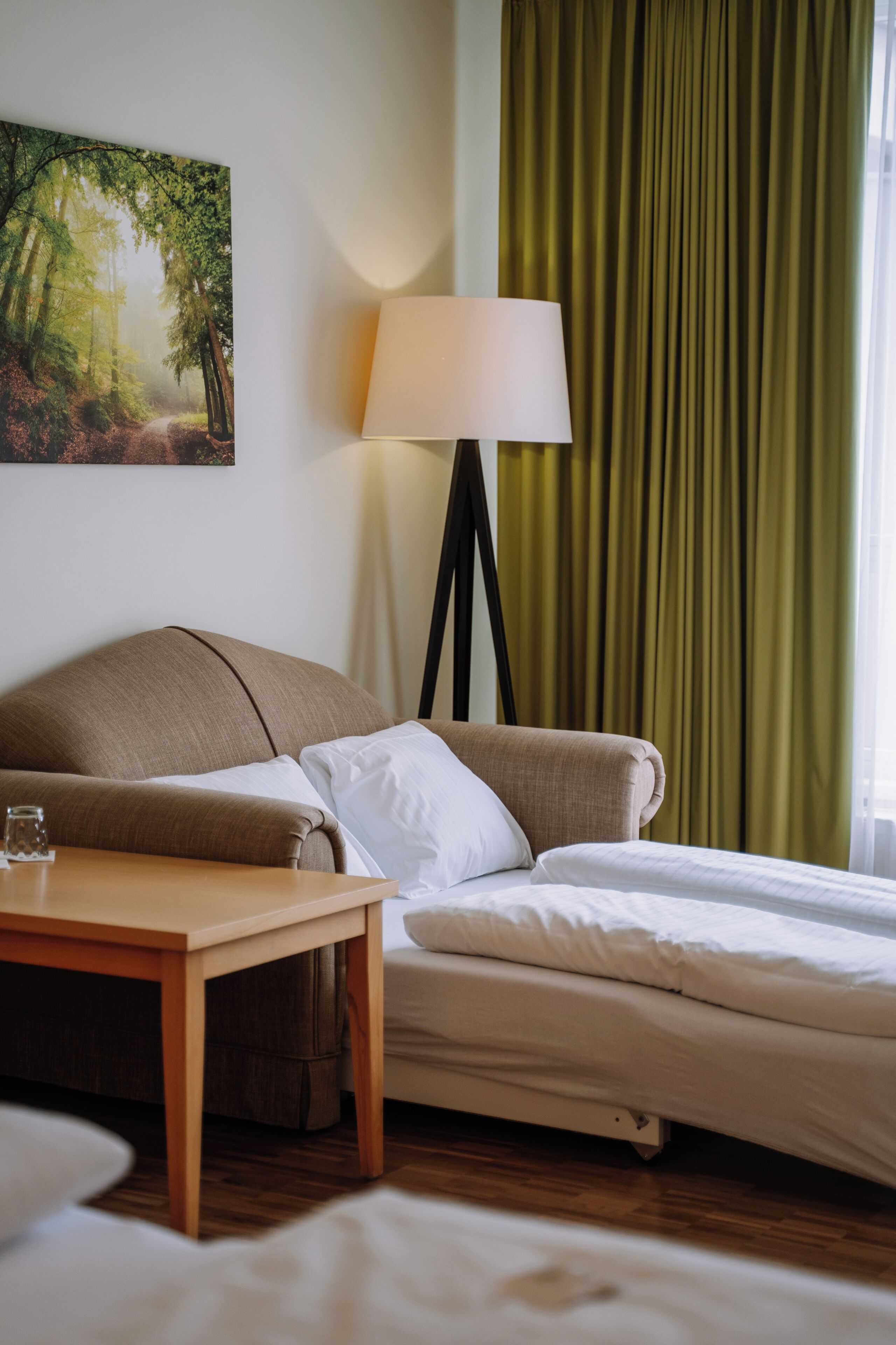 Habitación de hotel con cama, mesita de noche, lámpara y un cuadro de una escena boscosa.