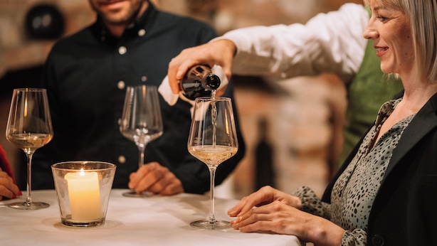 Une bougie dans un chandelier sur une table, un verre à vin et une personne qui verse une boisson dans un verre à vin.