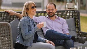 Un couple est assis sur un canapé d’extérieur en osier, avec une table devant eux, et ils tiennent chacun un verre à la main.