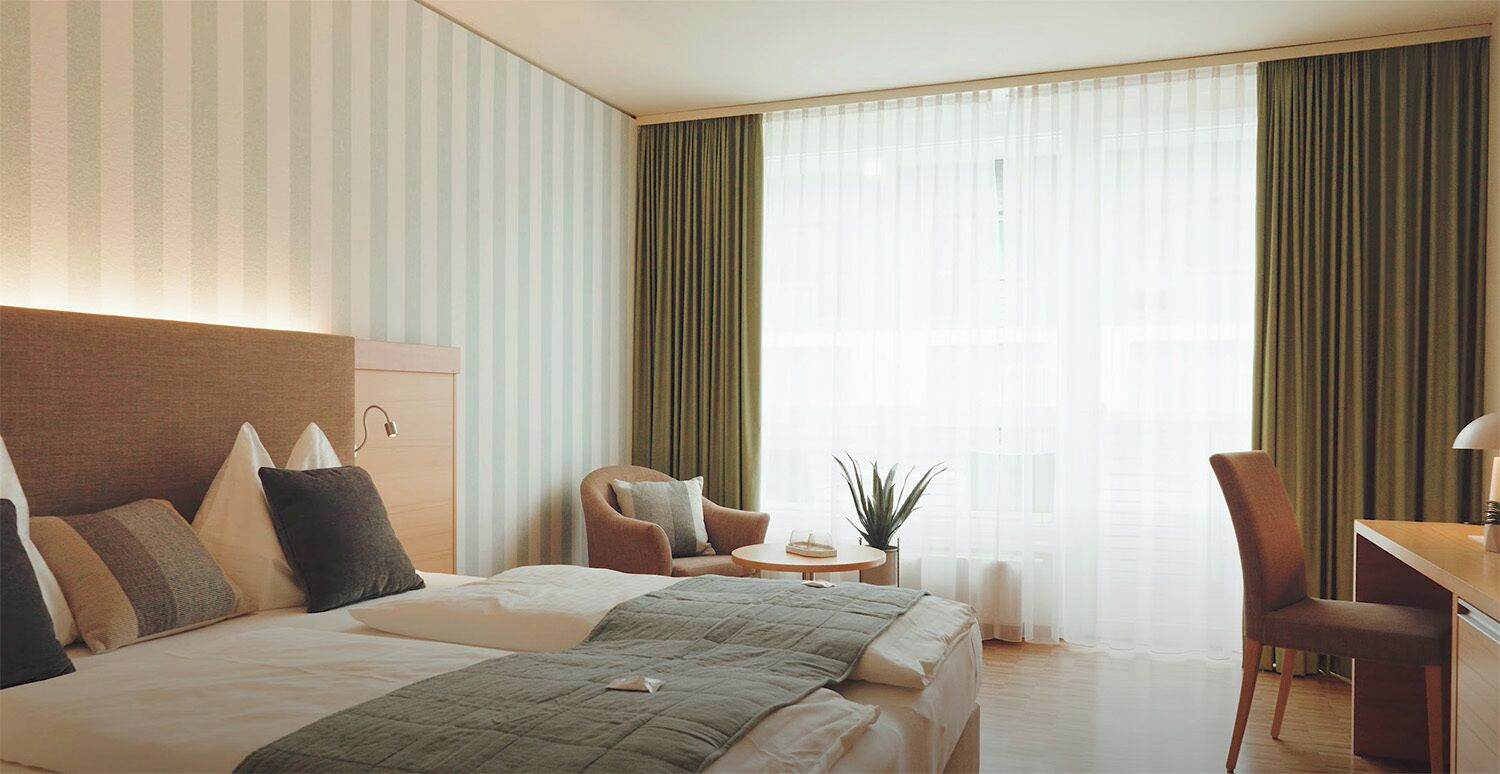Ein Hotelzimmer mit einem großen Bett, einem Schreibtisch, einem Stuhl, einem kleinen Tisch mit Pflanze und einem Fenster mit Vorhängen.