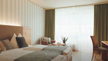 A hotel room with a large bed, a desk, a chair, a small table with a plant, and a window with curtains.