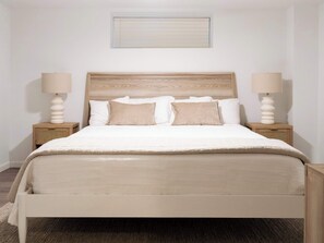 Un lit bien fait, avec une tête de lit en bois, deux tables de chevet avec des lampes, et un mur sobre avec une fenêtre au-dessus du lit.