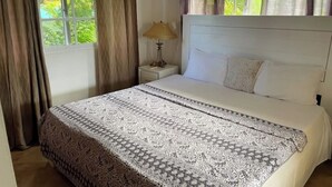 A neatly made bed with a patterned bedspread, a bedside lamp, and a window with curtains. - Villa Belle Amour (Sosúa)