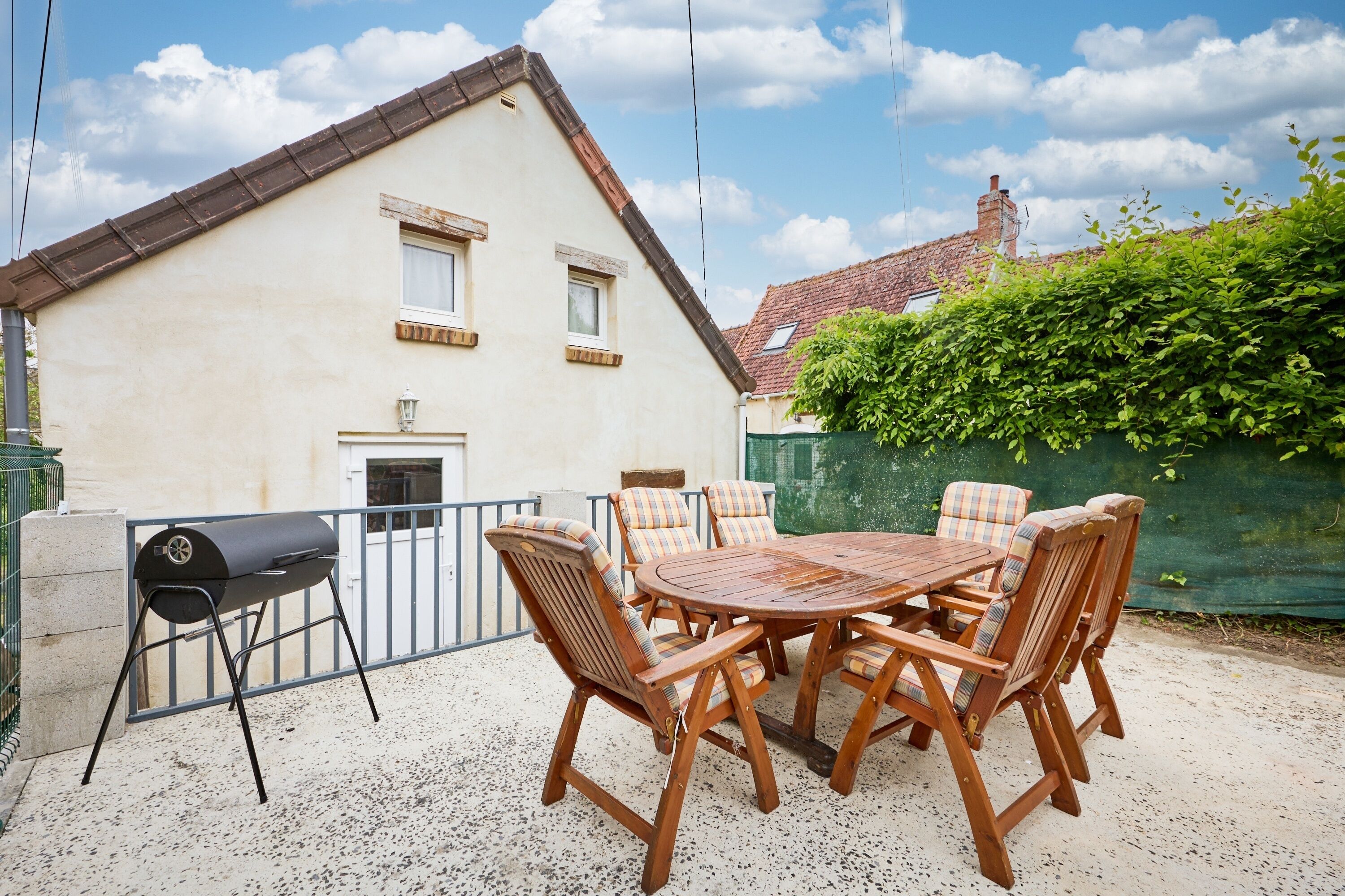 An outdoor patio with a round wooden table and six chairs, a barbecue grill, and a house in the background.