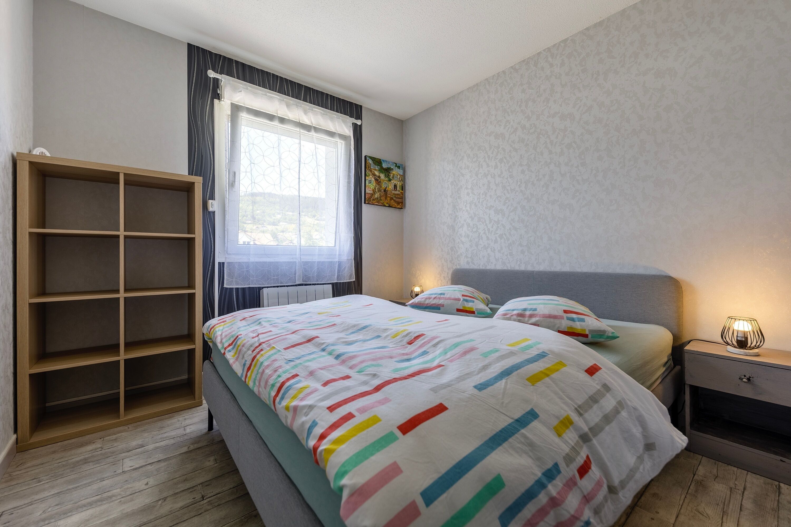 Une chambre à coucher comprenant un lit, une table de chevet, une armoire, une fenêtre avec des rideaux et un tableau accroché au mur.