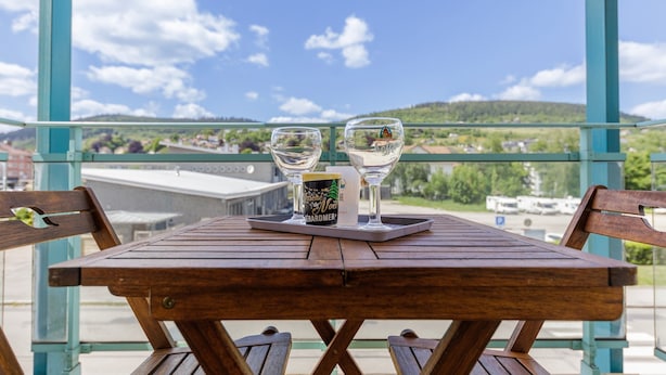 Una mesa de madera con dos vasos y una taza en un balcón con vista a un paisaje verde y edificios.