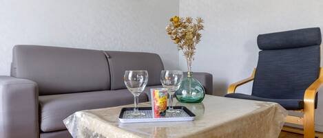 A living room with a sofa, a table with a tray, two glasses, a vase with dried flowers, and a chair.