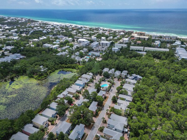 An aerial view of a residential area with houses, a swimming pool, and a beach in the background.