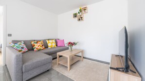 Un salon moderne avec un canapé gris, des coussins colorés, une table basse en bois, un téléviseur à écran plat posé sur un meuble en bois et une petite étagère ornée d’objets décoratifs.