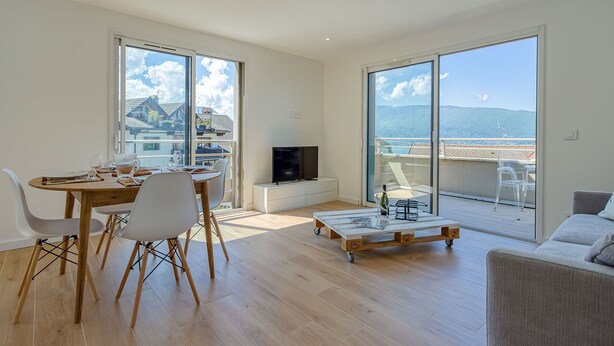 A modern living room with a dining area, large windows, a flat-screen TV, and a balcony with a view of the mountains. - Chez NiLo - 5min de la Plage (Veyrier-du-Lac)