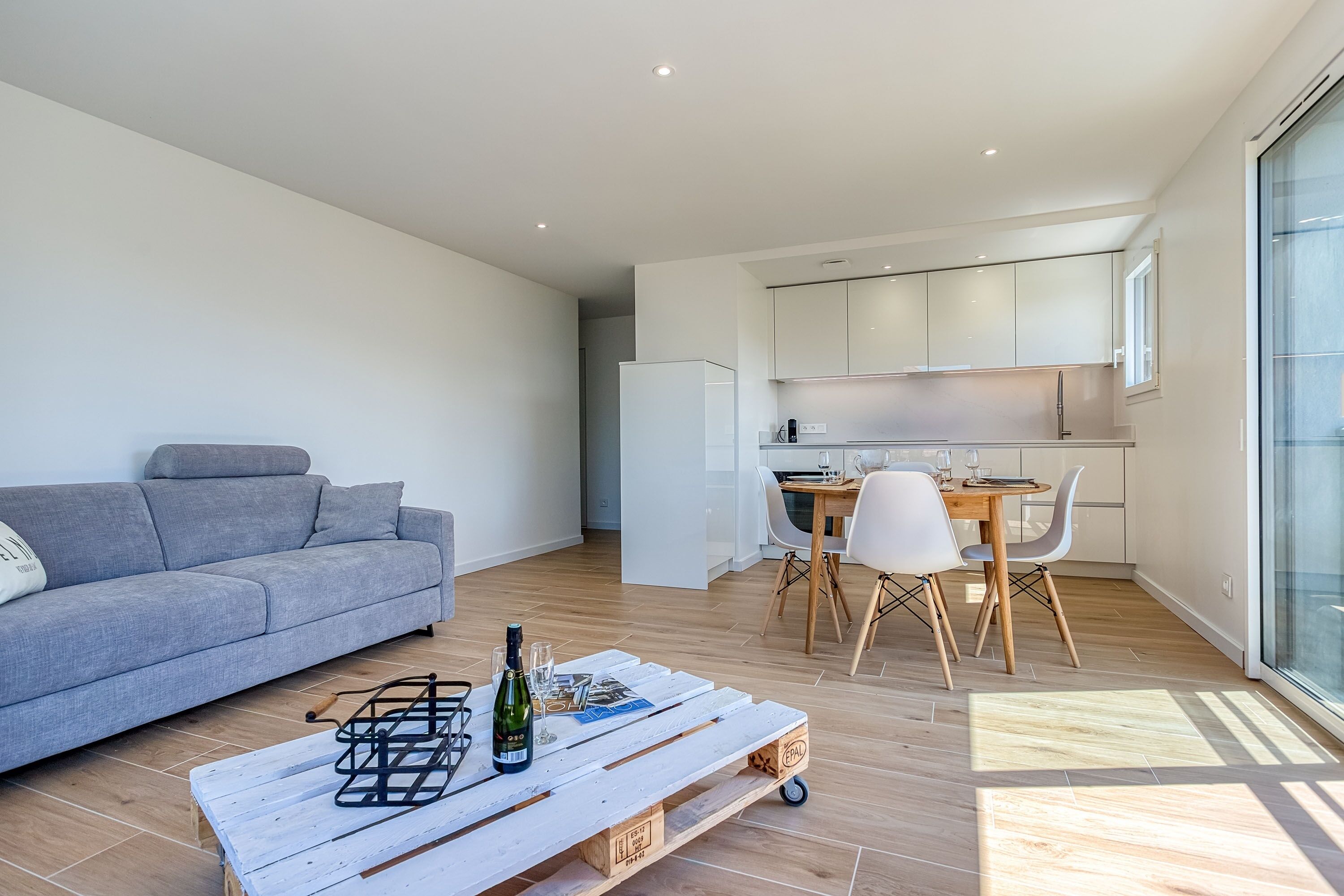 Una sala de estar moderna con un sofá gris, una mesa de centro de madera con una botella y vasos, y un comedor con mesa y sillas de madera.