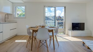 A modern kitchen with white cabinets, a dining table with chairs, a TV on a stand, and a large window with a view of buildings and trees. - Chez NiLo - 5min de la Plage (Veyrier-du-Lac)