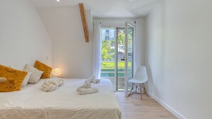 A bedroom with a bed, white and yellow pillows, a white chair, and a view of greenery through a glass door. - Chez NiLo - 5min de la Plage (Veyrier-du-Lac)
