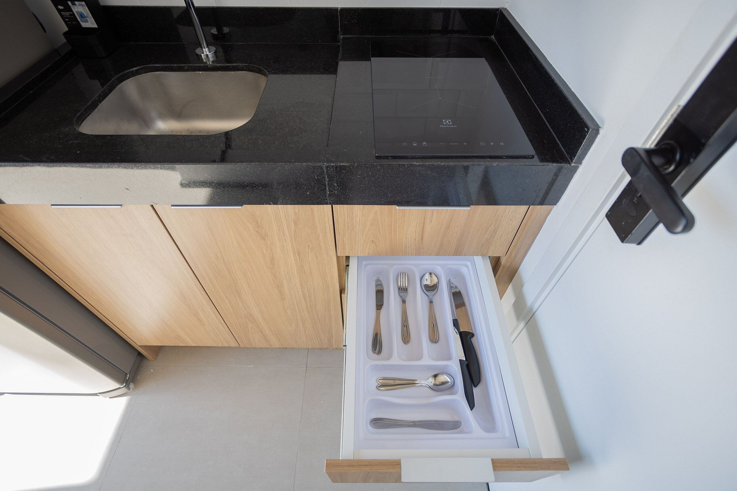 A compact kitchen with a black countertop, a stainless steel sink, and a drawer organizer with utensils.