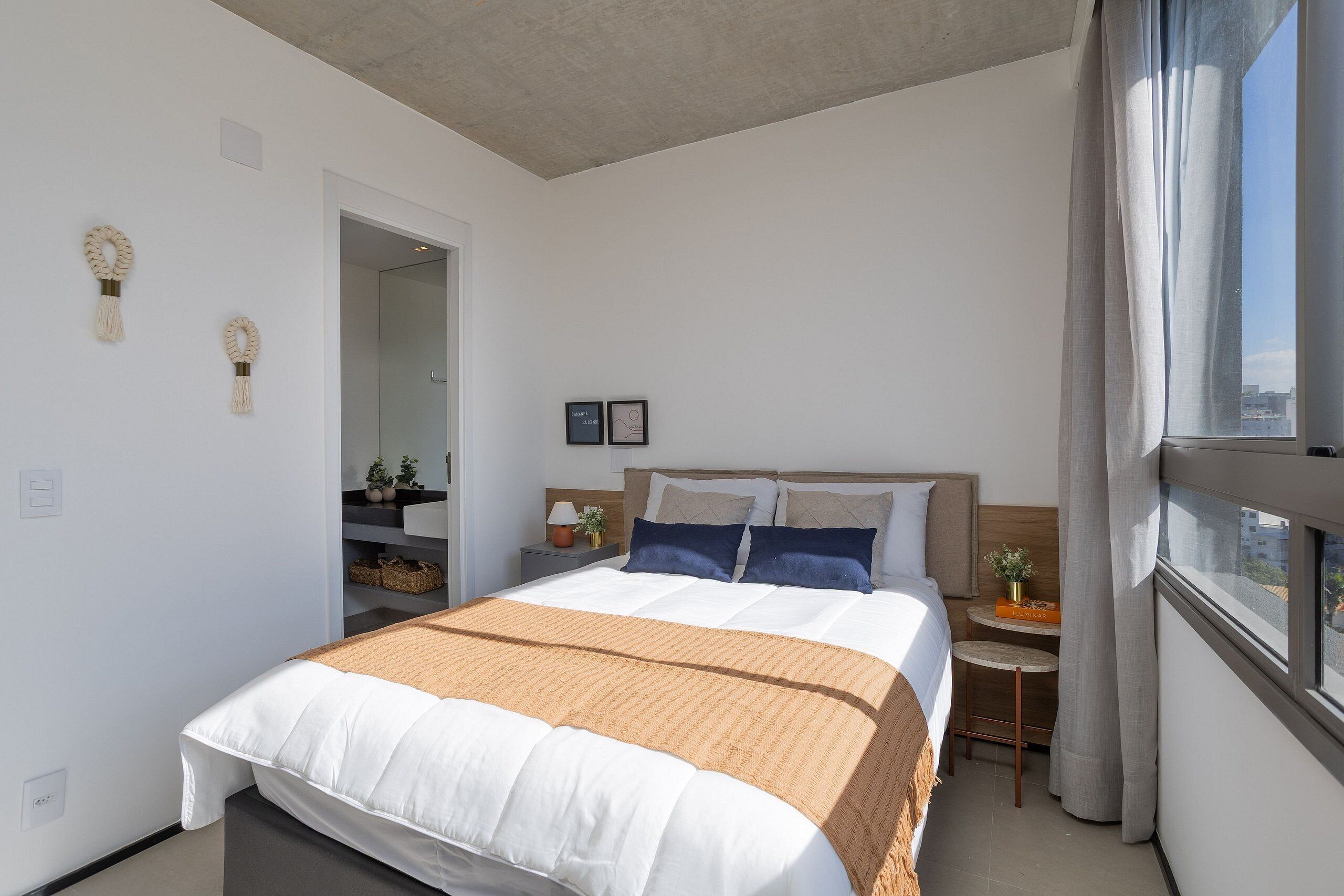 Moderni hotellihuone, jossa on suuri sänky, pieni yöpöytä, yövalaisin, ikkuna verhoilla ja näkymä kaupunkiin.