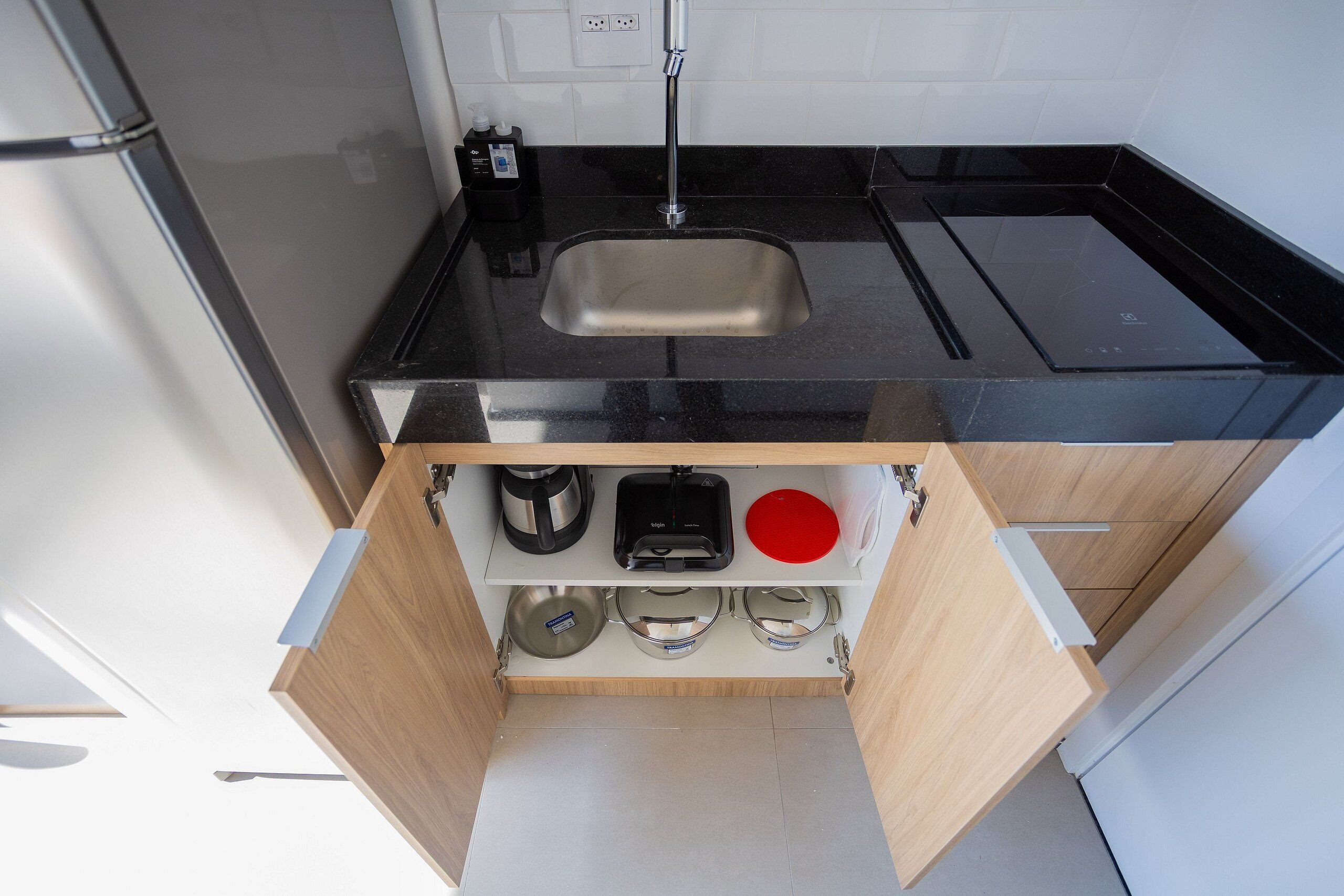 A kitchen with a black countertop, a stainless steel sink, and a wooden cabinet with a coffee maker, a red bowl, and three stainless steel containers.