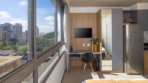 A modern hotel room with a large window, a desk with a chair, a flat-screen TV, a refrigerator, and a bed.