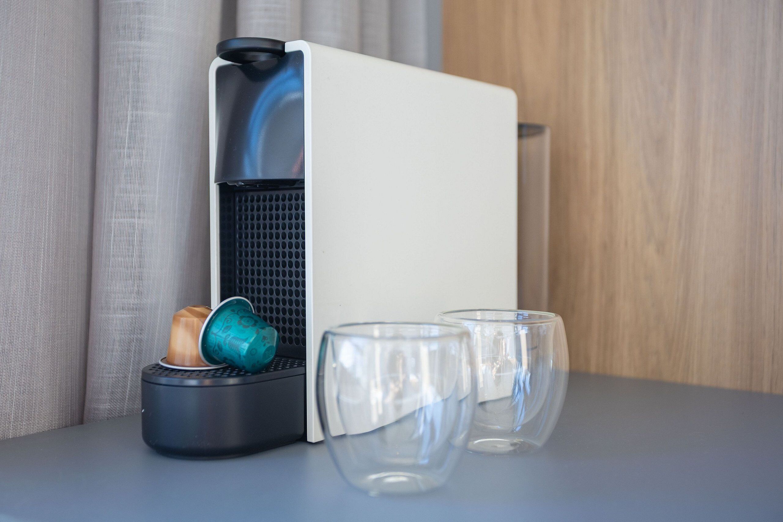 Una cafetera compacta con depósito de agua y portavasos, dos copas de vino vacías sobre una mesa.