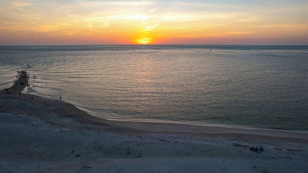 Une plage où l’on peut admirer le coucher de soleil sur l’océan, une rive sablonneuse et quelques personnes qui se promènent.