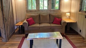 A cozy room with a wooden floor, a sofa with red cushions, a table, and a window with a view of trees. - Cherry Treesort “Traci’s Treasure” (Rowan County)