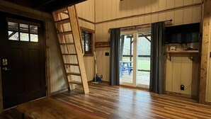 A room with a wooden floor, a wooden bench, a ladder, a door, and a window with curtains. - Cherry Treesort “Traci’s Treasure” (Rowan County)