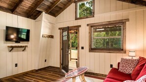 A cozy room with a wooden floor, a red sofa, a small round table, a TV mounted on the wall, and a window with blinds. - Cherry Treesort "The Tolson" (Rowan County)