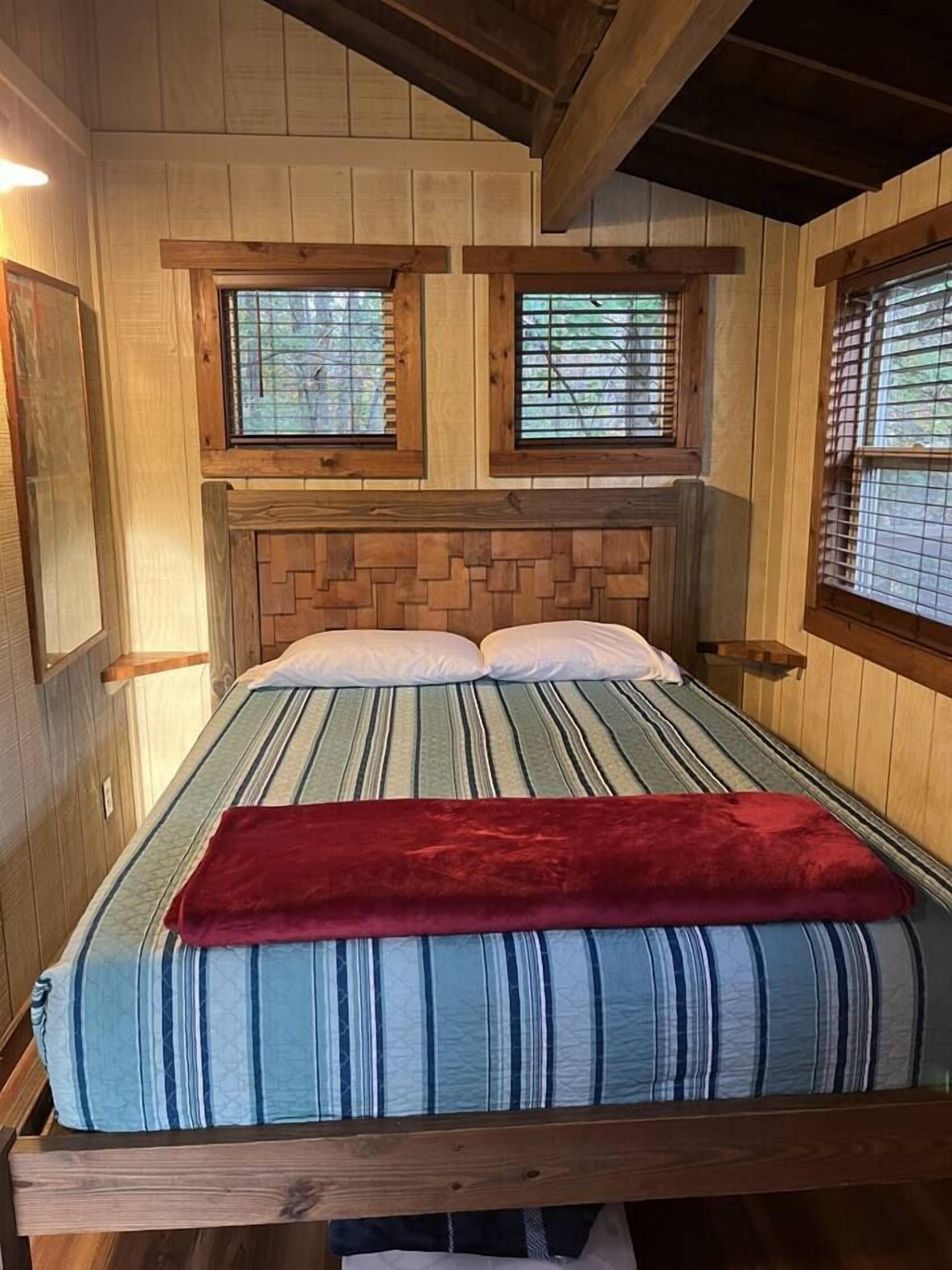Una habitación con paneles de madera, una cama, dos ventanas con persianas y una colcha roja sobre la cama.